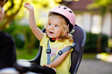 Portrait of little toddler girl with security helmet on the head sitting in bike seat of parents. Boy on bicycle on background. Safe and child protection concept. Family and weekend activity trip.