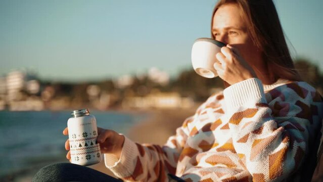 Young beautiful woman in cozy sweater and sunglasses filling cup of coffee from thermos while relaxing on winter seaside sand beach. Cute attractive girl enjoying hot tea taking sunbathe near ocean.