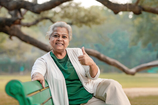 Indian Senior Woman Sitting On Bench And Showing Thumps Up At Park.