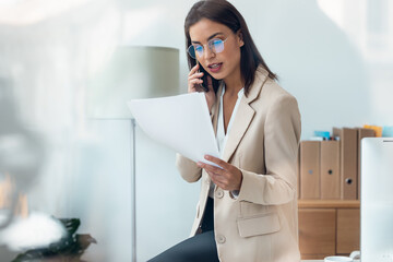 Elegant beautiful businesswoman talking with smartphone while reading some documents sitting on the desk at office