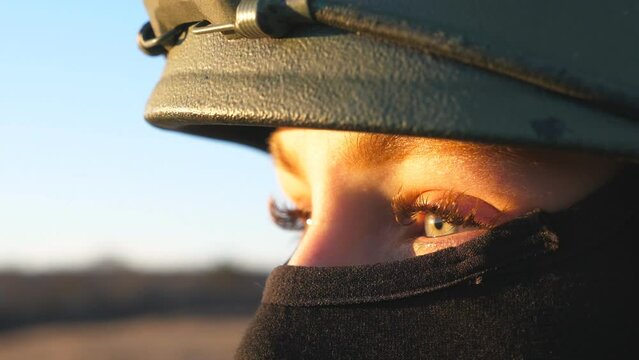 Close up green eyes of female ukrainian army soldier looking at sunset against blue-yellow flag. Sad sight of military woman in helmet and balaclava at countryside. Resistance to russian invasion