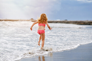 Happy Child, Little Preschool Girl in Swimmsuit Running And Jumping In The Waves During Summer Vacation On Exotic Tropical Beach. Family Journey On Ocean Coast.