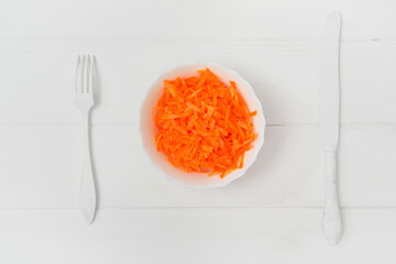 Flat lay, composition, freshly grated carrot in a plate. On the wooden table, a knife and fork. White background. Light key.