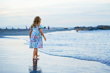 Happy Child, Little Preschool Girl in Dress Running And Jumping In The Waves During Summer Vacation On Exotic Tropical Beach by Sunset. Family Journey On Ocean Coast.