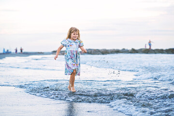 Happy Child, Little Preschool Girl in Dress Running And Jumping In The Waves During Summer Vacation On Exotic Tropical Beach by Sunset. Family Journey On Ocean Coast.