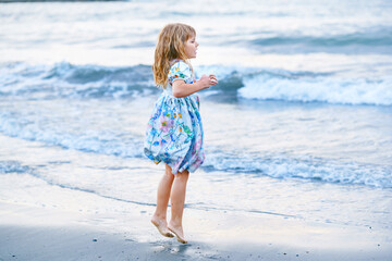 Happy Child, Little Preschool Girl in Dress Running And Jumping In The Waves During Summer Vacation On Exotic Tropical Beach by Sunset. Family Journey On Ocean Coast.