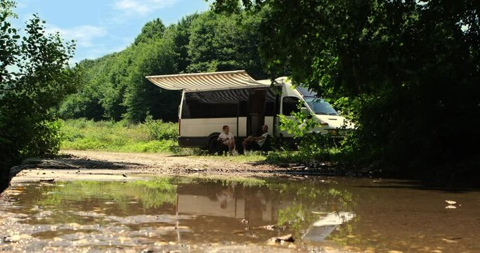 In the  of the forest, a couple living the caravan life sits in their chairs by the stream, savoring the beauty of nature 