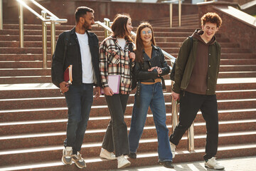 Walking, holding notepads. Four young students in casual clothes are together outdoors