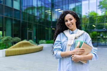 Portrait of a young beautiful Hispanic female student, woman outside a university campus smiling and looking at the camera, holding workbooks, books and textbooks for studying.