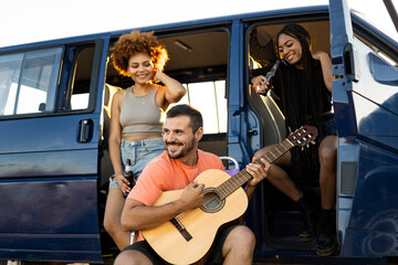 A group of 3 young people from all walks of life are having fun in a camper van parked on a mountain. The man plays a flamenco guitar while the African girls drink beer. Travel with a camper.