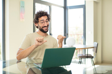 young adult bearded man with a laptop smiling cheerfully and casually pointing to copy space on the side, feeling happy and satisfied