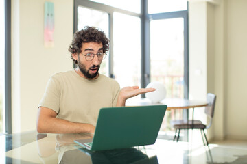 young adult bearded man with a laptop looking surprised and shocked, with jaw dropped holding an object with an open hand on the side