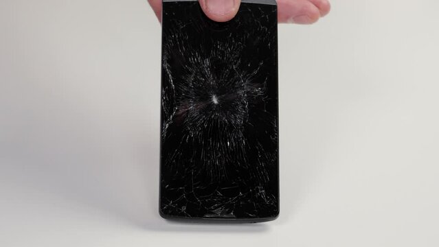 A person picking up a broken mobile phone with a shattered touchscreen from a white floor close up