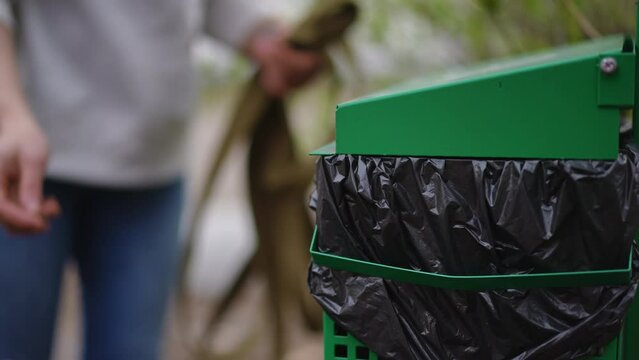 Close-up special trash can in park with unrecognizable woman throwing away dog poop leaving. Closeup female Caucasian hand placing garbage in bin outdoors