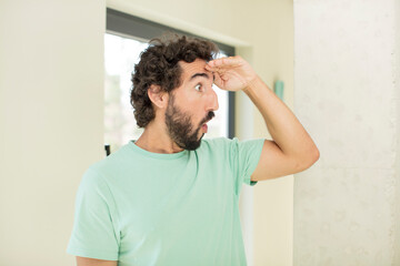 young crazy bearded man looking bewildered and astonished, with hand over forehead looking far away, watching or searching