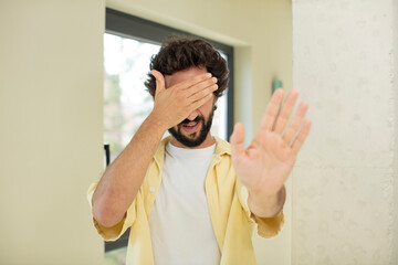 young crazy bearded man covering face with hand and putting other hand up front to stop camera,...
