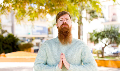 red hair bearded man feeling worried, hopeful and religious, praying faithfully with palms pressed,...