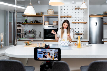 Young woman vlogger broadcasting live video online teaching cooking food in kitchen at home, recording and streaming on smartphone. Cook lessons from home studio for young people.