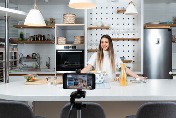 Young woman vlogger broadcasting live video online teaching cooking food in kitchen at home, recording and streaming on smartphone. Cook lessons from home studio for young people. - obrazy, fototapety, plakaty