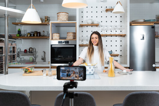 Young woman vlogger broadcasting live video online teaching cooking food in kitchen at home, recording and streaming on smartphone. Cook lessons from home studio for young people.
