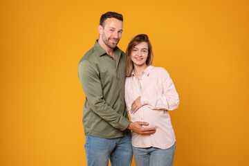 Happy young european couple awaiting baby, parents-to-be enjoy tender moment man hugging pregnant wife and touching belly posing over yellow background