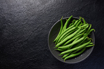Green pods of raw asparagus beans in a bowl on a black stone background. Top view with copy space.