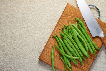 Wooden cutting board with pods of raw asparagus beans and a knife on a light marble background with copy space. Top view.