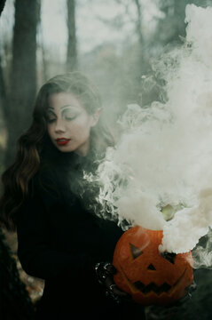 A young woman in a Gothic gloomy image of a witch surrounded by smoke, a pumpkin in her hands. Halloween costume. Vertical photo.