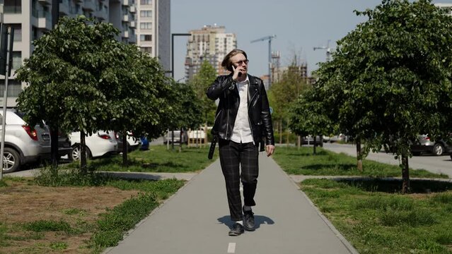 A business man in a leather jacket and plaid trousers walks to a residential area. Talking on the phone, white shirt. Slow motion.