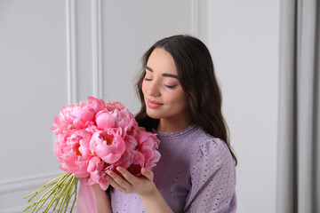Beautiful young woman with bouquet of pink peonies near white wall indoors
