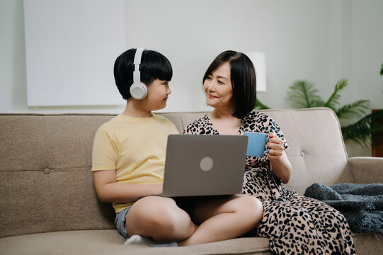 Homeschool Asian Young Little Boy Learning Online And Does Homework By Using Computer And Tablet With Mother Help, Teach And Encourage. Son Smile To Study At Home Together