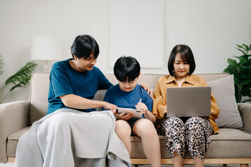 Asian parents and kids laugh use devices together sit on sofa, tech addicted family with children hold laptop phone digital tablet having fun at home