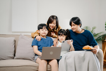 Asian parents and kids laugh use devices together sit on sofa, tech addicted family with children hold laptop phone digital tablet having fun at home