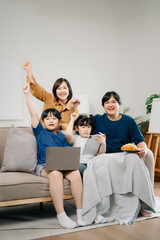 Asian parents and kids laugh use devices together sit on sofa, tech addicted family with children hold laptop phone digital tablet having fun at home