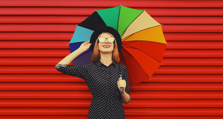 Autumn portrait of stylish happy smiling young woman holding colorful umbrella wearing black round hat on red background