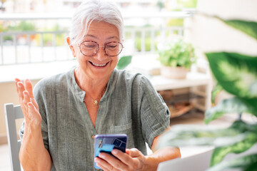 Video call concept. Happy senior woman with eyeglasses having video call on mobile phone sitting outdoor on terrace, elderly lady using modern technology and wireless connection