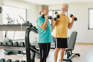 Happy old senior couple in sportswear exercising with dumbbells in gym. Old man and woman doing stretching exercise to stay fit