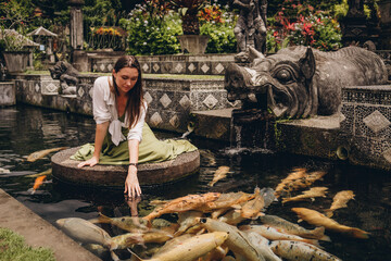 Beautiful girl sitting and admiring pond koi fishes. Smiling girl feeding carps in Ubud Water Palace