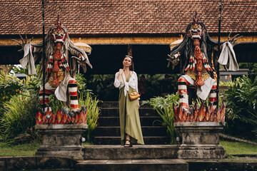 Beautiful girl between two statues with barong mask. Religious hindu ritual temple with colorful...