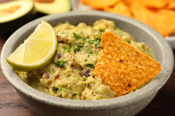 Bowl of delicious guacamole, lime and nachos chips on wooden table, closeup