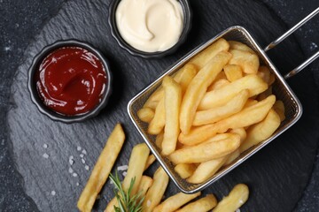 Tasty French fries, ketchup, rosemary and mayonnaise on black textured table, top view