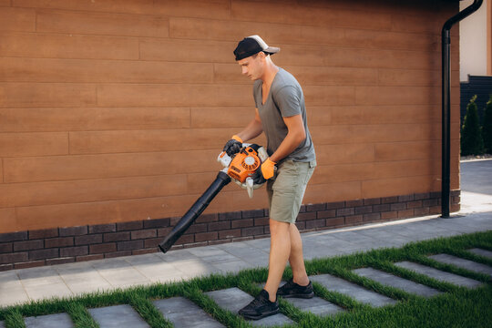 Low angle view of caucasian worker in overalls, white cap and gloves removing dry leaves from lower with hand blower. Gardener taking take of back yard during summer season.