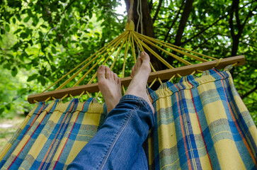 Bare female legs, dressed in jeans, on a checkered hammock, outdoor recreation in the forest, among the greenery