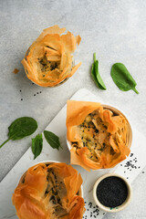 Filo pies with soft feta cheese and spinach in ceramic molds on old grey table background. Filo portions pies. Small Baked Spanakopita pies. Top view. Copy space. Traditional Greek cuisine.
