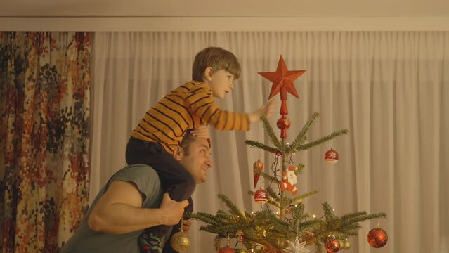 Man rises piggyback son to arrange the star decoration on top of Christmas fir tree