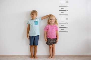 Little children, blond boy and girl, measuring height against wall in room - obrazy, fototapety, plakaty