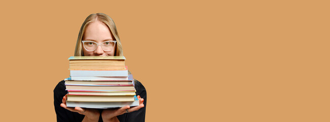 Caucasian female student in glasses holds a lot of books in her hands on a beige background. Book Lovers Day. Long banner. - obrazy, fototapety, plakaty
