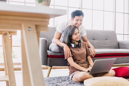 Happy Asian Couple Shopping Online With Laptop Computer At Home. Lovely Couple Making Purchases Online Or Booking In Beautiful Living Room. Buying Ordering Products On The Internet.