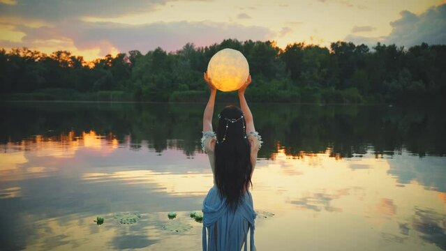 Fantasy woman and moon, holding glowing planet in hands raises to sky prays. Girl fairy tale elf river nymph wet dress, stands in water dark lake summer nature. Lady queen night back rear view. art 4k