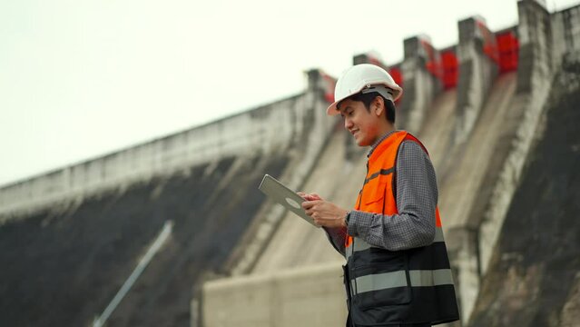 Confident asian maintenance engineer man inspection with tablet in construction site dam with hydroelectric power plant and irrigation. Engineer man wearing safety hat at project big building.